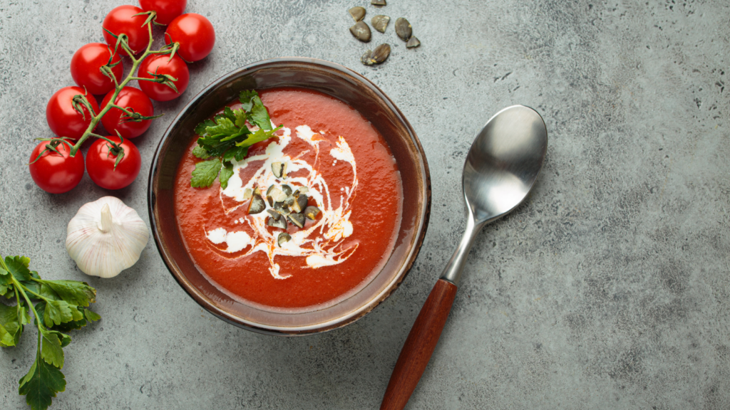 Bowl of gazpacho topped with herbs and seeds, surrounded by tomatoes, garlic, and parsley on a stone surface.